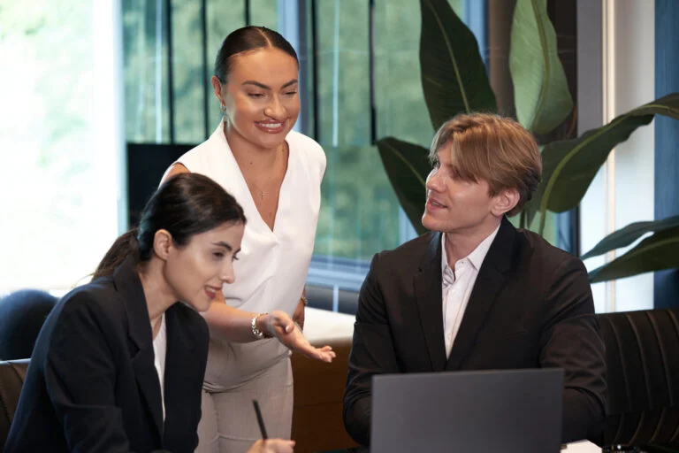 Three people in business attire have a discussion around a laptop in a modern office setting featuring large windows and plants, ideal for those seeking a private meeting room Oakville or flexible day-use office options. - Business Lounge Oakville