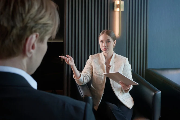A woman holding a clipboard speaks to a man in an office setting, both seated on black chairs, discussing meeting room membership Oakville for flexible workspace solutions. - Business Lounge Oakville