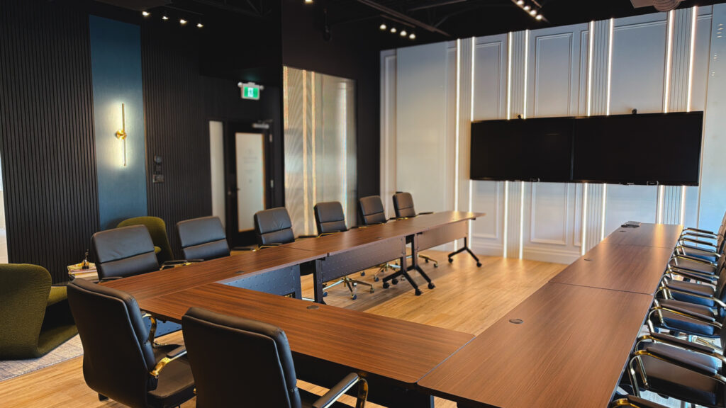 Modern conference room with U-shaped wooden tables, black office chairs, wall-mounted screens, and wood flooring, lit by ceiling lights—ideal as a quarterly offsite meeting room in Oakville. - Business Lounge Oakville
