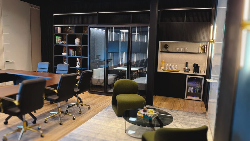 Modern office lounge with green chairs, a round coffee table, conference table, bookshelves, and a kitchenette with beverages and coffee machine—ideal as a team training room Oakville. Glass-walled meeting room in the background. - Business Lounge Oakville